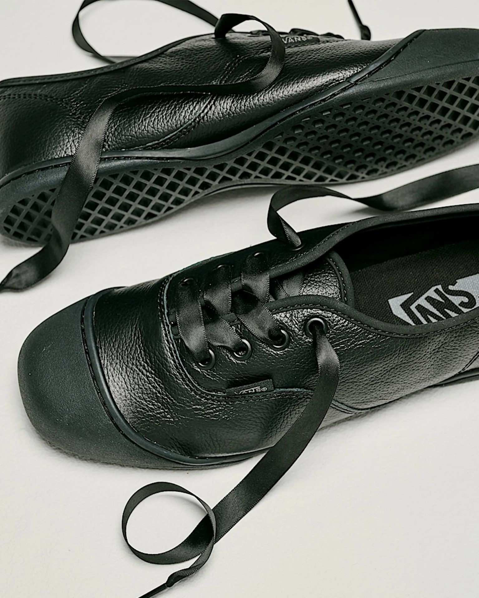 Pair of black leather Vans sneakers with satin ribbon laces on a light surface.