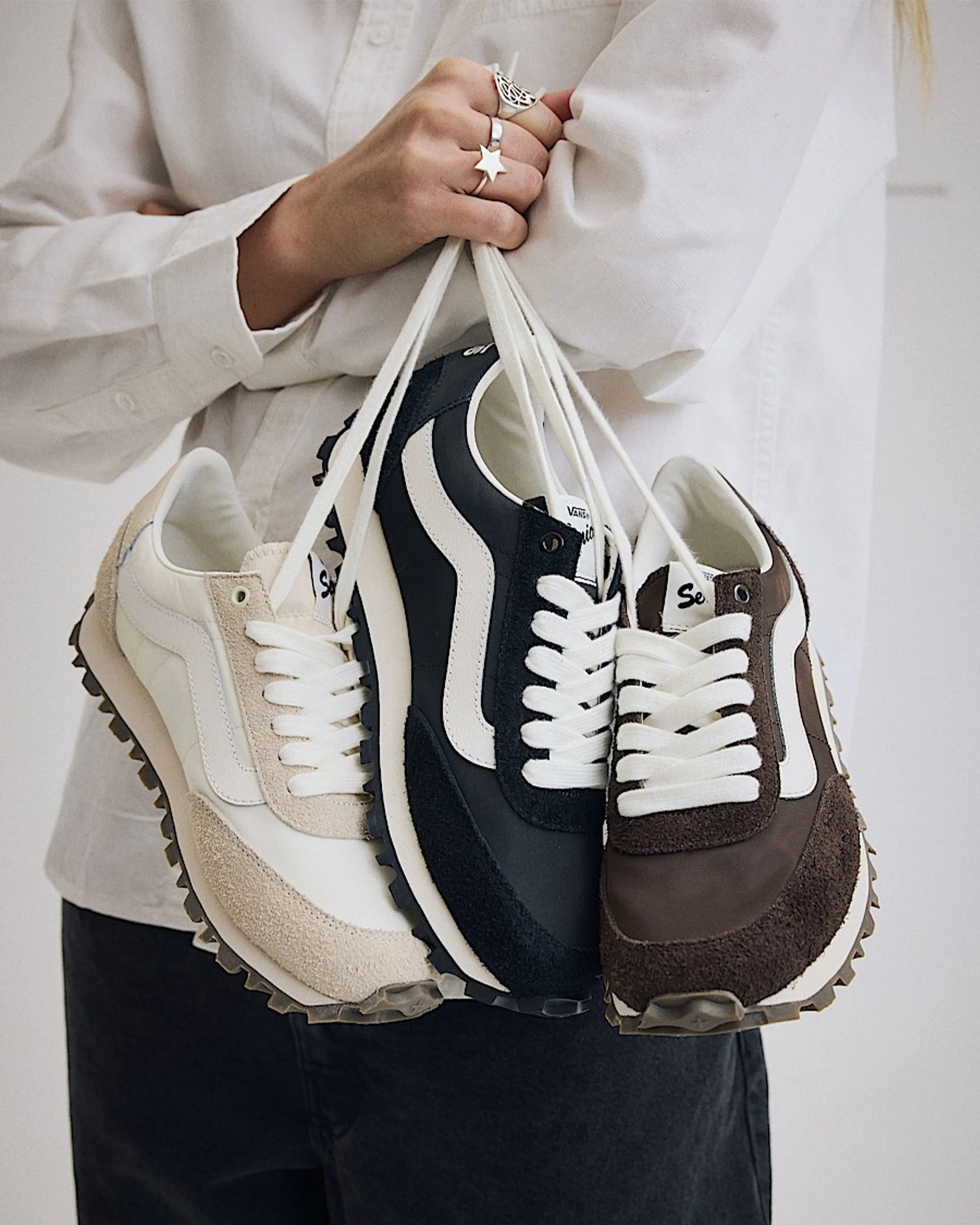 Three pairs of Vans sneakers in beige, black, and brown with white laces held by their laces in a person's hand.
