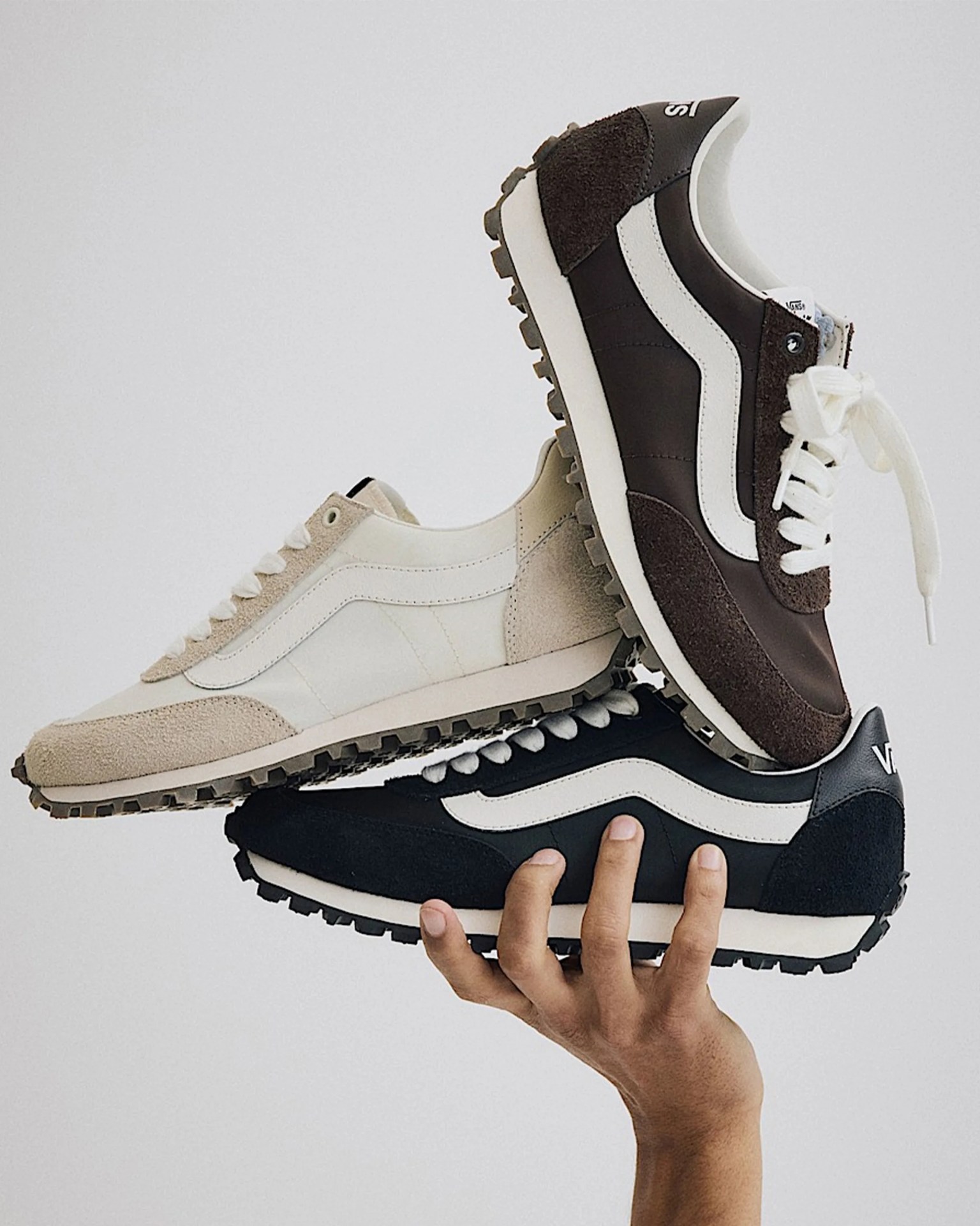 Three Vans sneakers in beige, brown, and black stacked and held in a hand against a plain background.