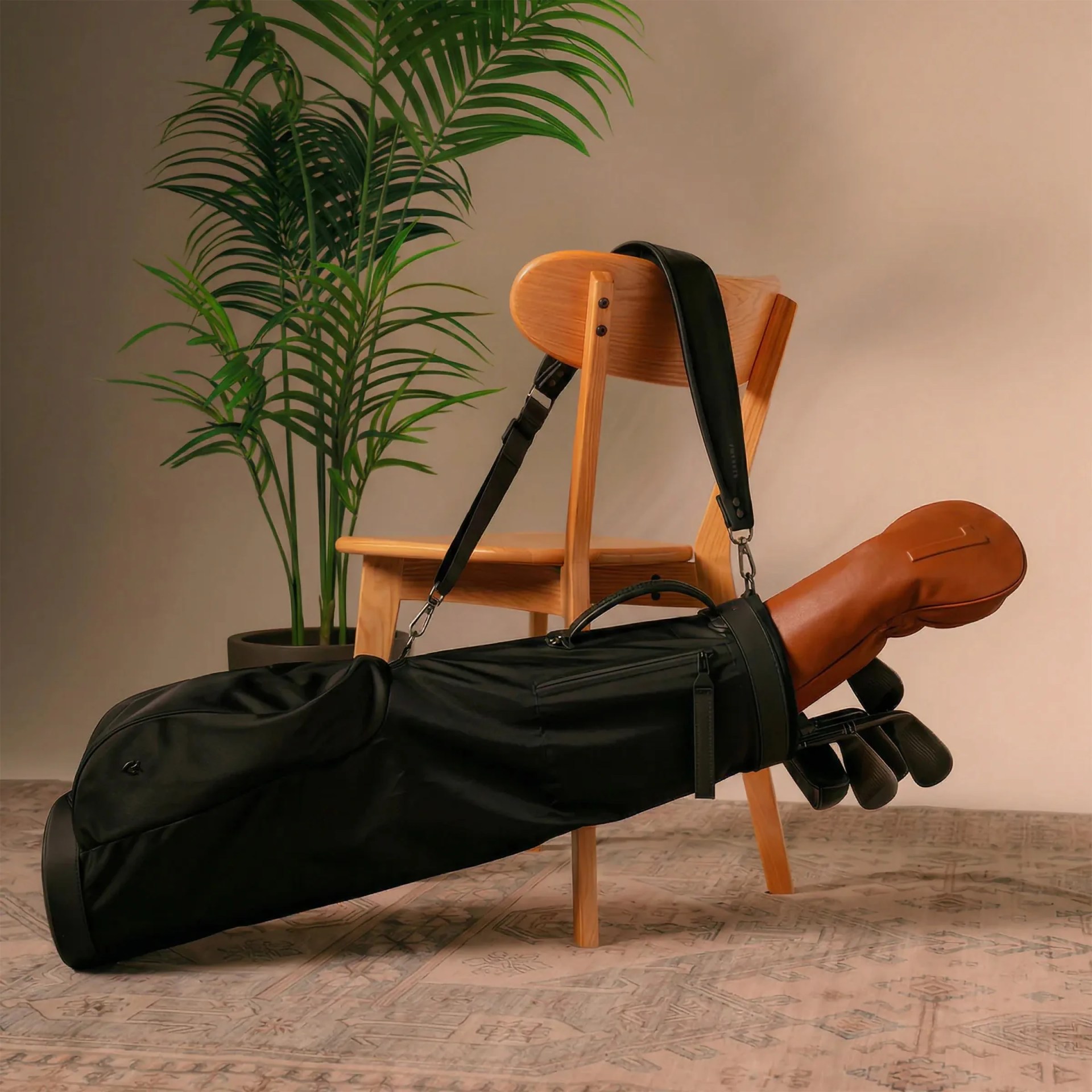 Black golf bag with clubs and a brown leather headcover resting on a wooden chair beside a potted green plant.