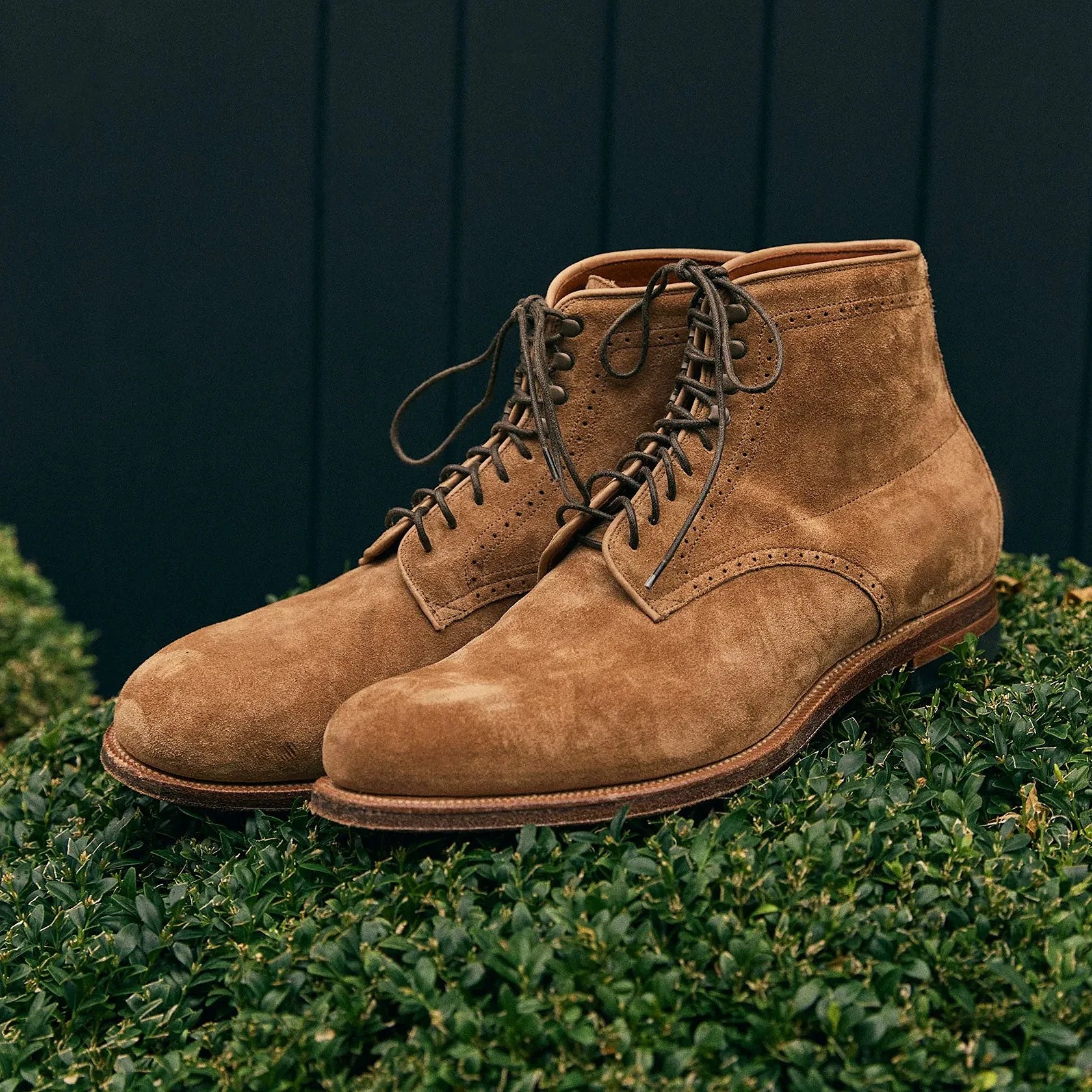 Pair of tan suede lace-up boots resting on green shrubbery with a dark vertical panel background.