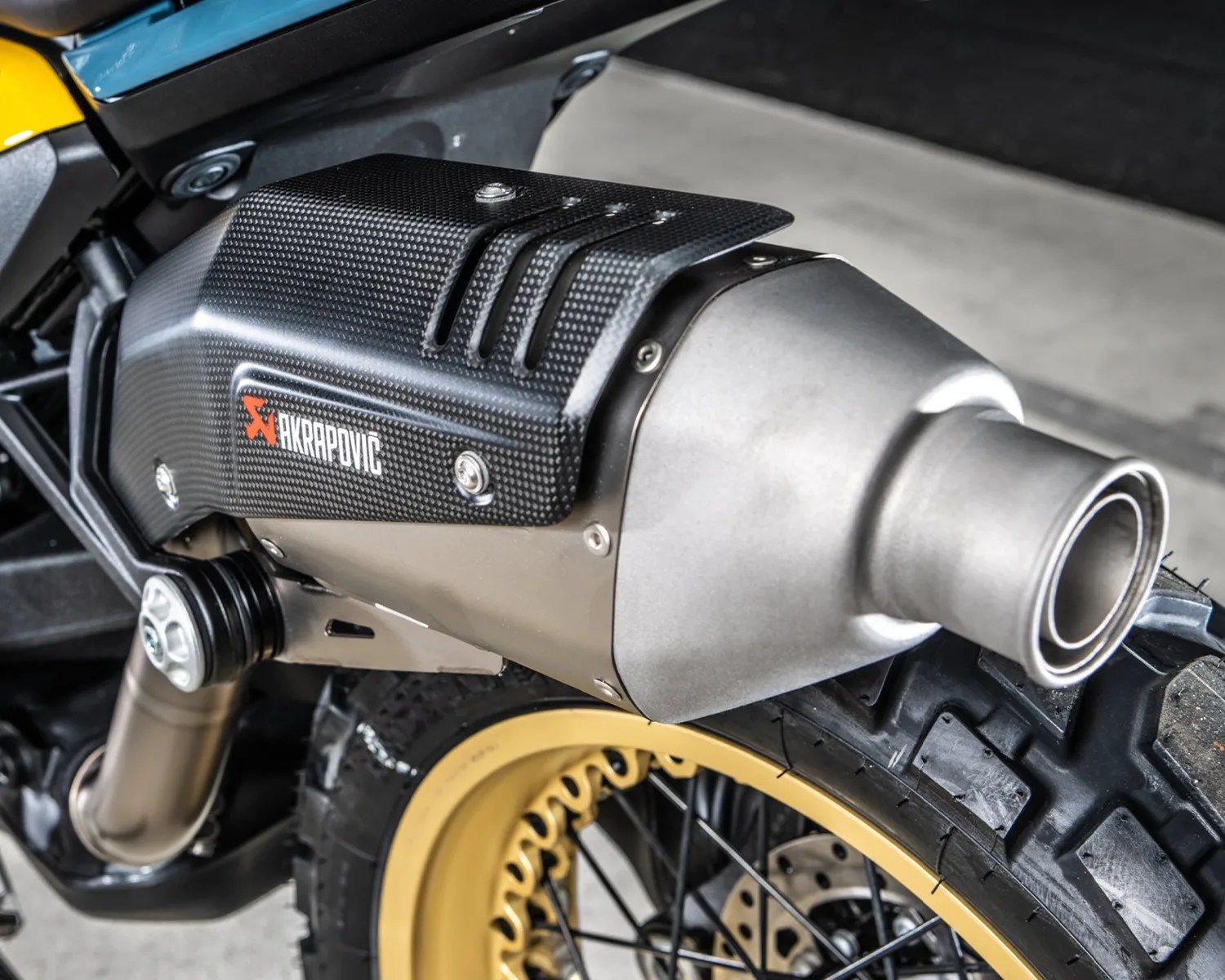 Close-up of a motorcycle exhaust with a black carbon fiber Akrapovic heat shield and a silver metal muffler above a yellow-rimmed tire.