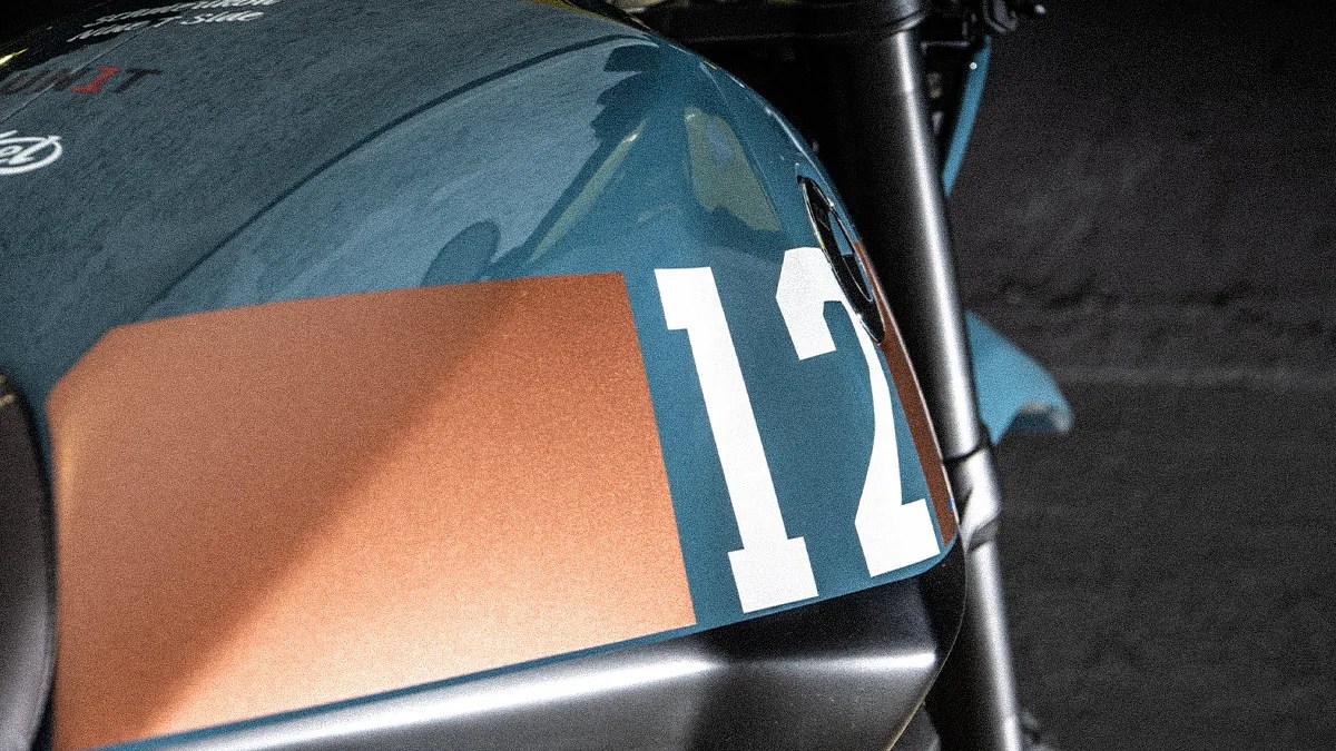 Close-up of a blue motorcycle fuel tank with a large white number 12 and a copper-colored rectangular decal.