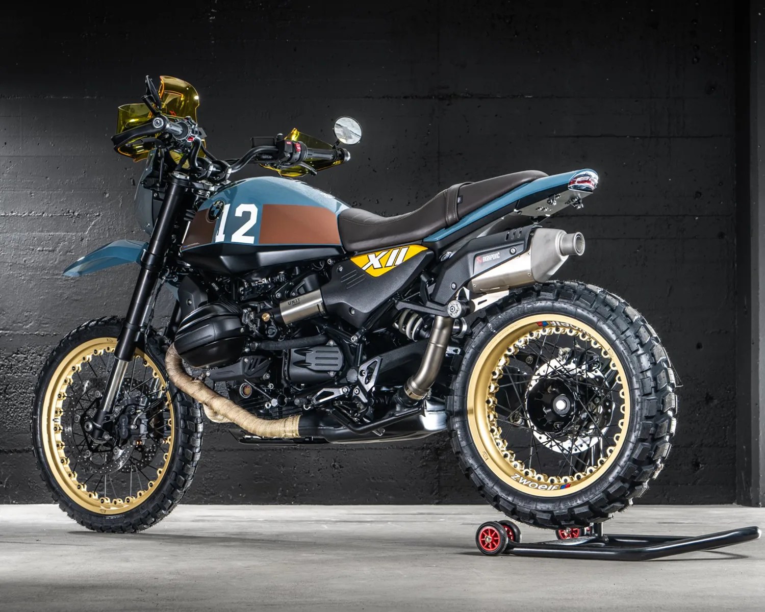 Blue and brown custom scrambler motorcycle with gold rims and knobby tires on a rear stand.