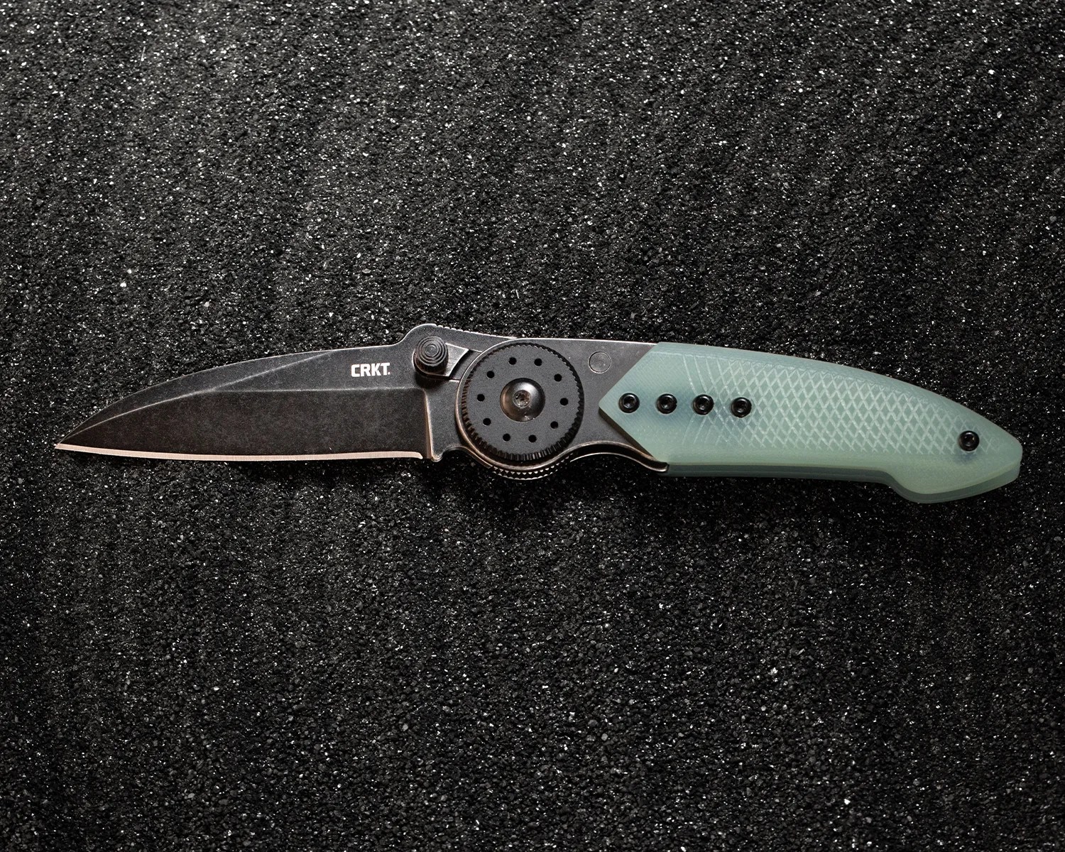 Black folding knife with a stonewashed blade and translucent green textured handle on a black textured surface.
