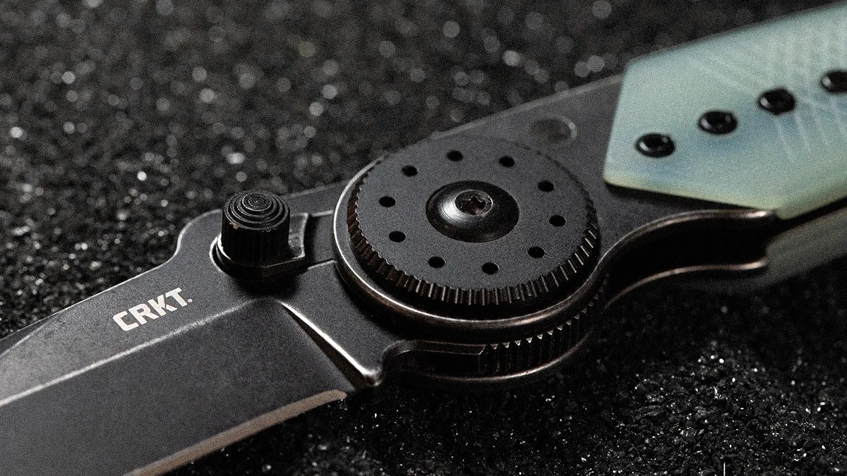 Close-up of a black CRKT folding knife with a textured thumb stud and circular pivot on a dark textured surface.