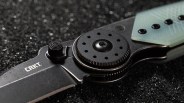 Close-up of a black CRKT folding knife with a textured thumb stud and circular pivot on a dark textured surface.