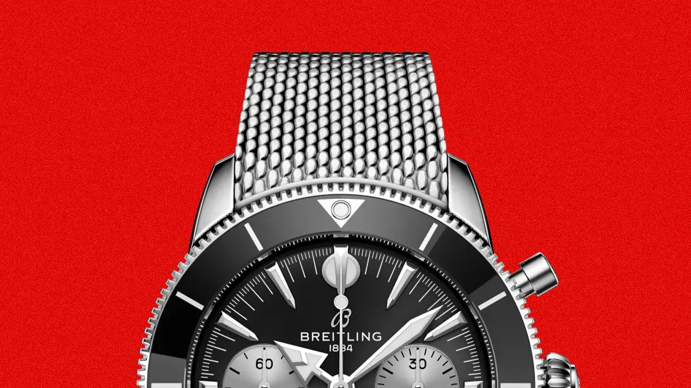 Breitling chronograph watch with black dial, silver subdials, and stainless steel mesh bracelet on red background.