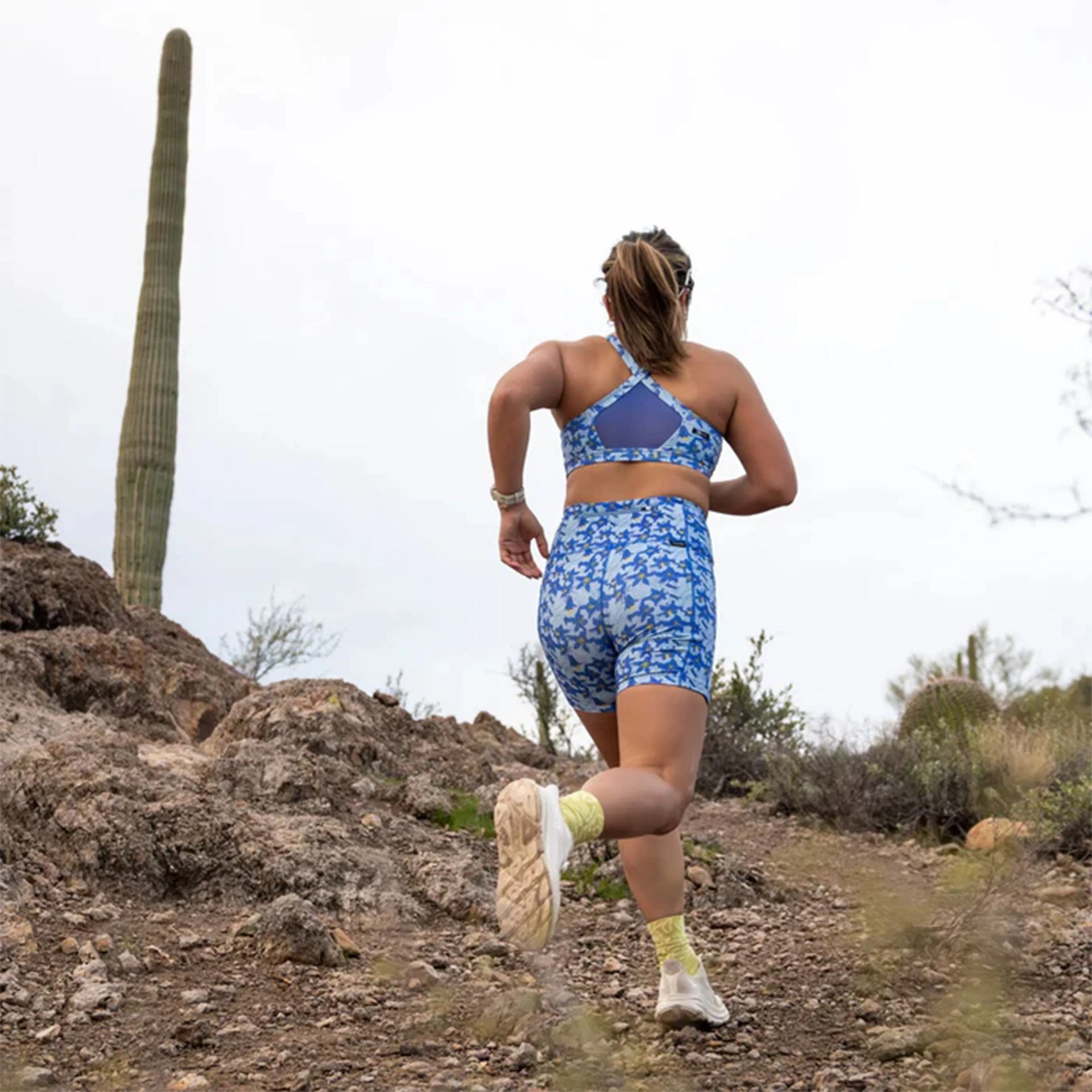 Person running on a rocky trail wearing blue patterned sports bra and shorts with yellow socks and white shoes.