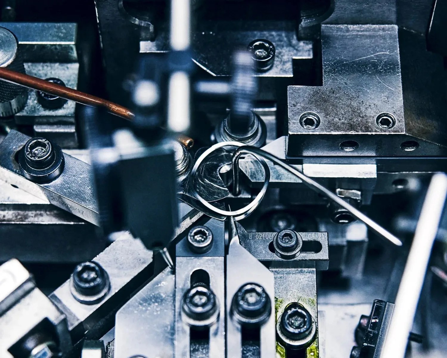 Close-up of a metal spring being shaped and assembled within a complex industrial machine with screws and metal components.