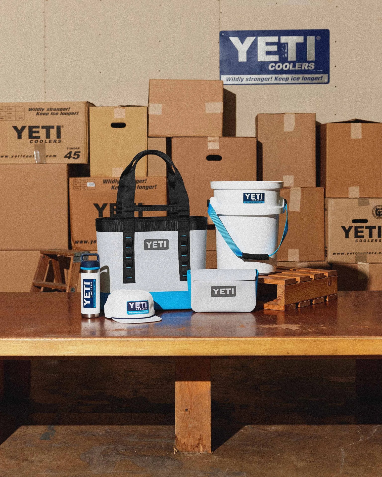 White and blue YETI branded cooler bag, bucket, water bottle, cap, and pouch displayed on a wooden table with stacked cardboard boxes in the background.