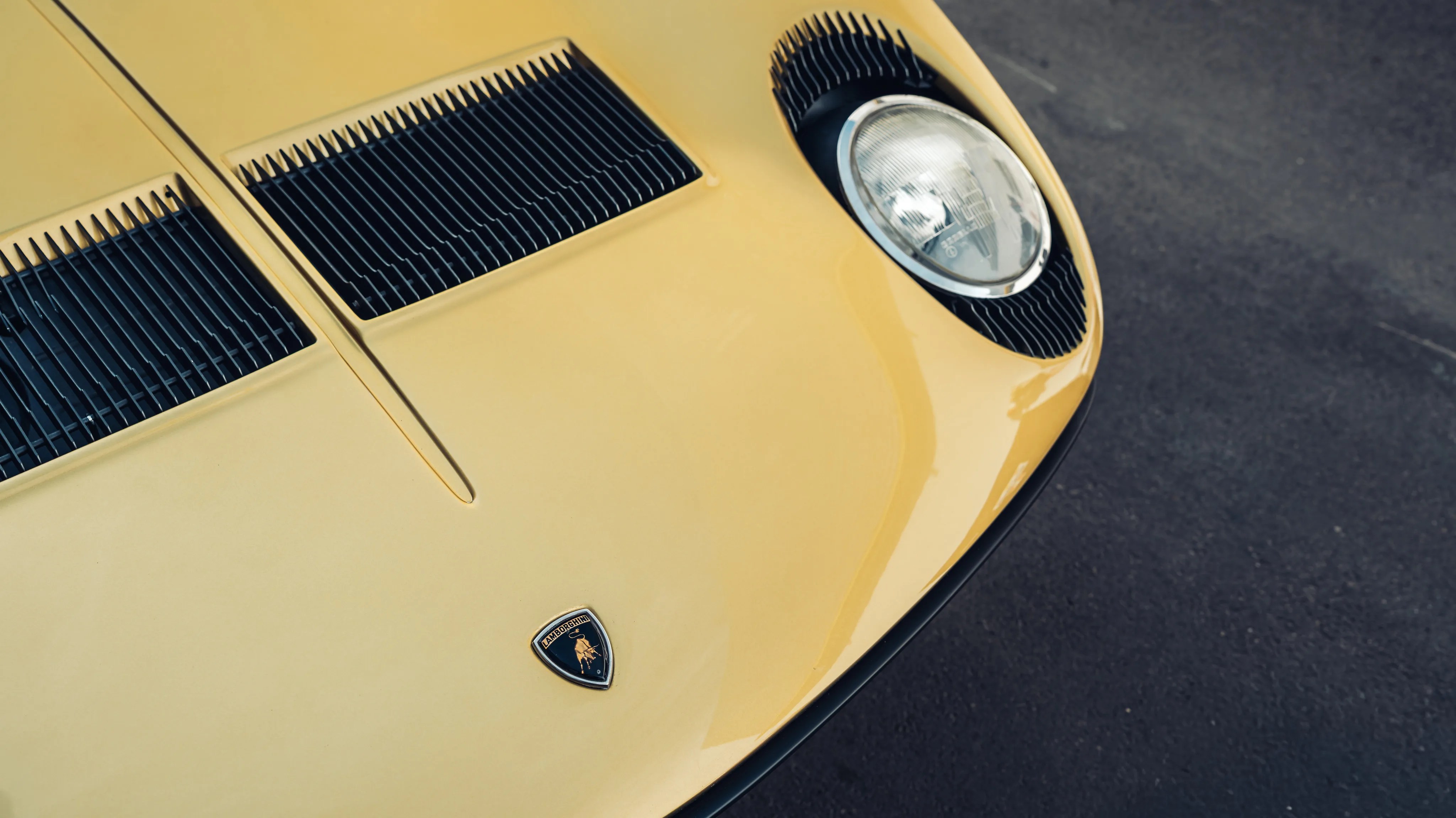 Close-up of a yellow Lamborghini hood with black vents and a round headlight on a dark asphalt surface.
