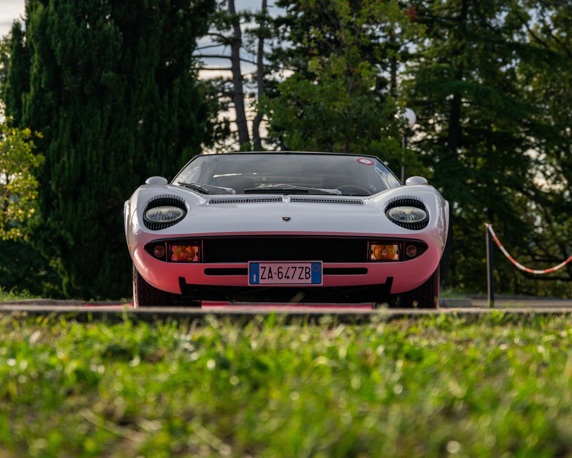 White classic Lamborghini Miura sports car front view with eyelashes on headlights, parked outdoors with greenery.