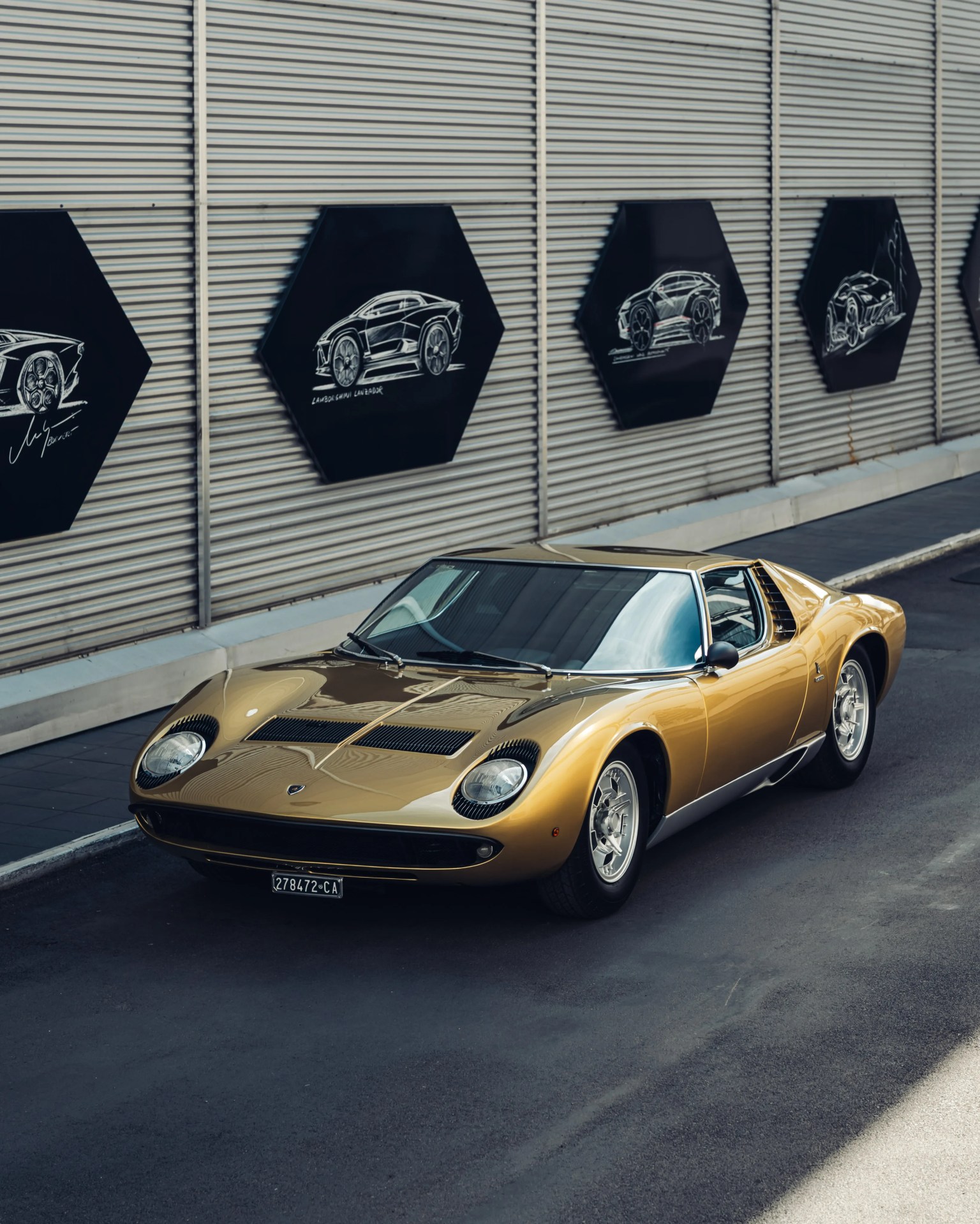 Gold classic Lamborghini Miura sports car parked on a street with hexagonal car sketches on a corrugated metal wall behind.