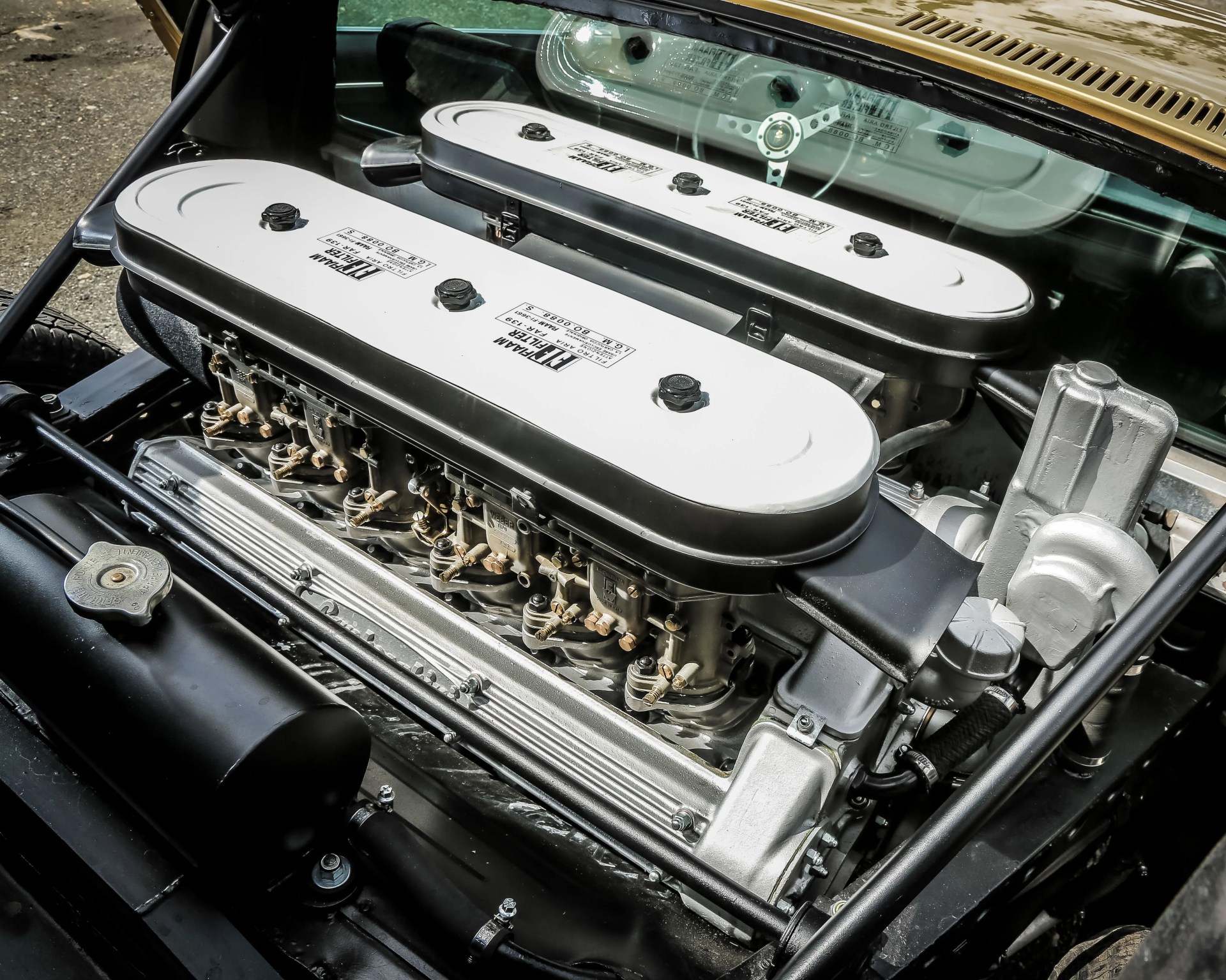 Close-up of a classic car engine with dual white and black air filters labeled "Ferrari" and visible carburetors.