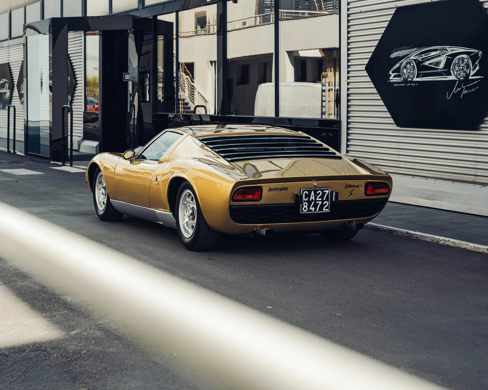 Gold Lamborghini Miura S sports car parked on a street near a modern building with reflective windows.