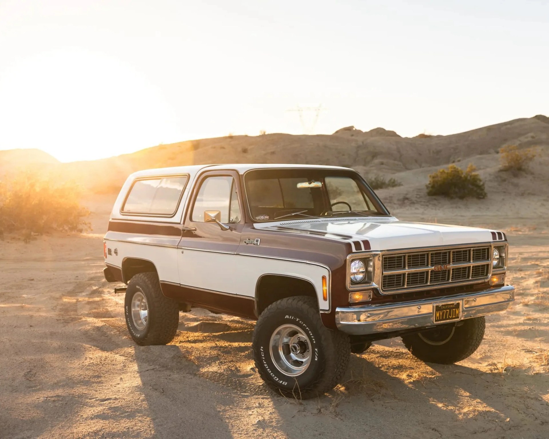 Two-tone brown and white vintage GMC SUV with off-road tires parked on sandy terrain at sunset.