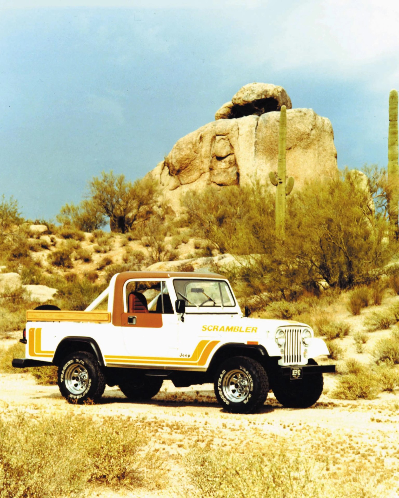 White Jeep Scrambler pickup truck with orange accents parked in a desert landscape with cacti and large rocks.