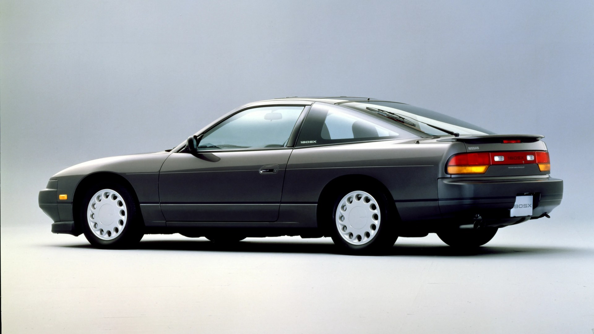 Dark gray Nissan 180SX coupe with white wheels shown in profile on a plain background.