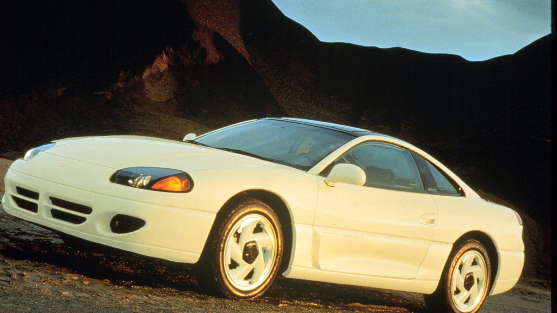 White two-door sports coupe with pop-up headlights parked on rocky terrain at sunset.
