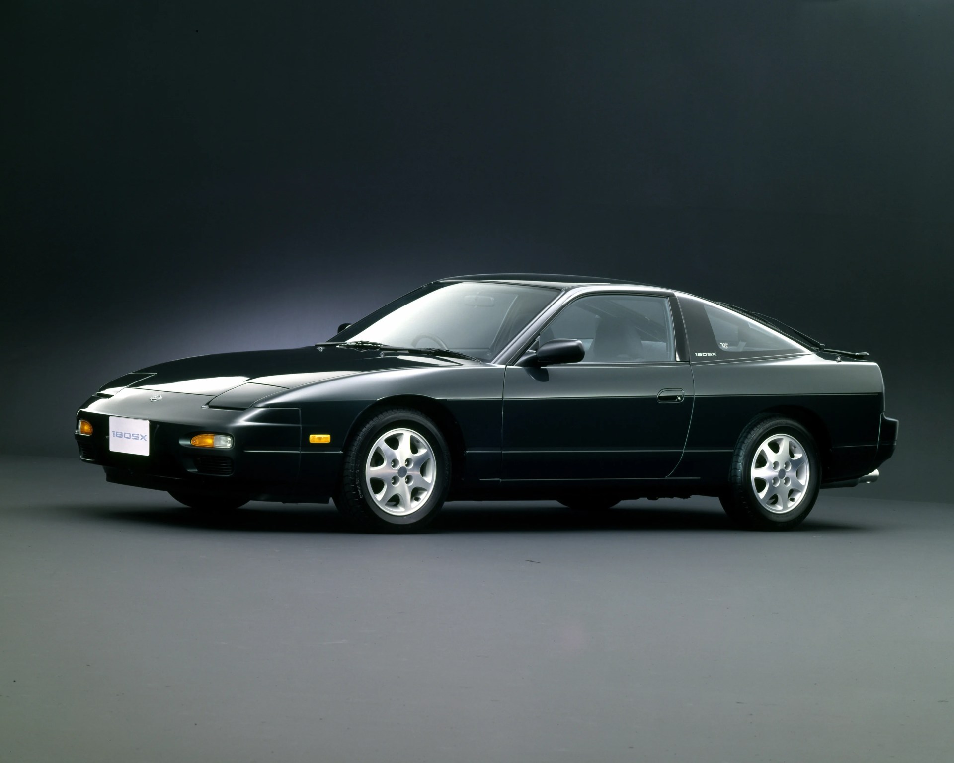 Black Nissan 180SX coupe with pop-up headlights and silver alloy wheels on a gray studio floor.