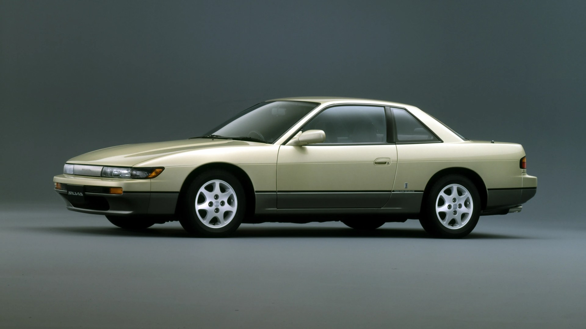 Beige and gray two-tone Nissan Silvia coupe with five-spoke silver wheels on a gray studio background.