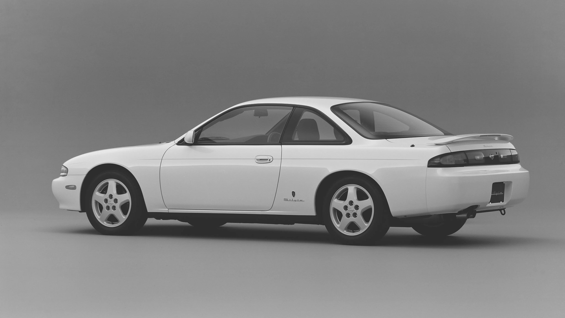 White Nissan Silvia coupe with rear spoiler and five-spoke alloy wheels on a gray background.