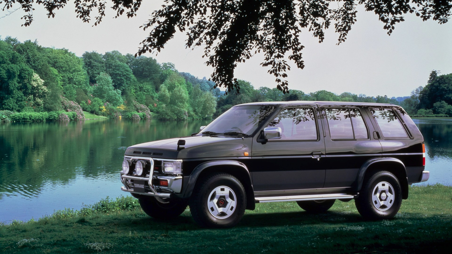 Black Nissan Terrano SUV parked on grass near a lake with trees in the background.