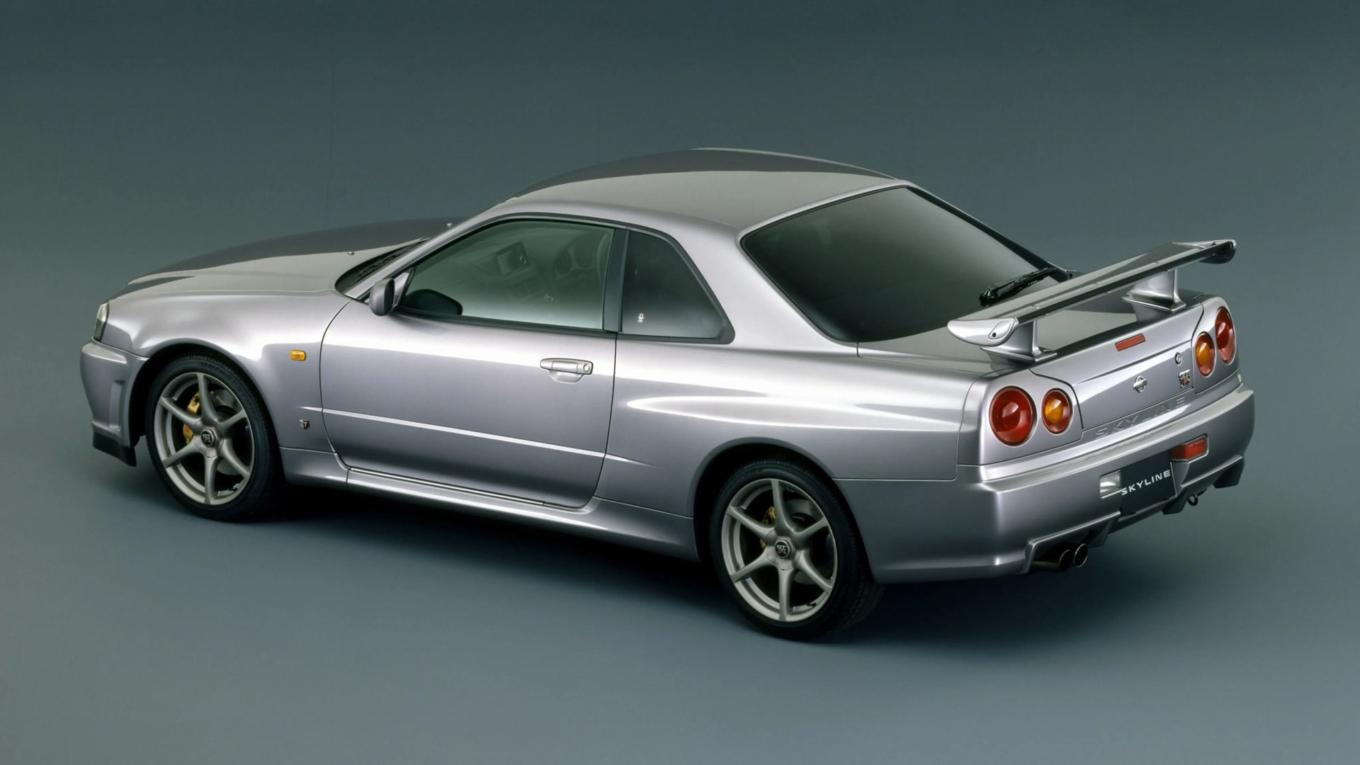 Silver Nissan Skyline GT-R R34 coupe with rear spoiler on a gray studio background.
