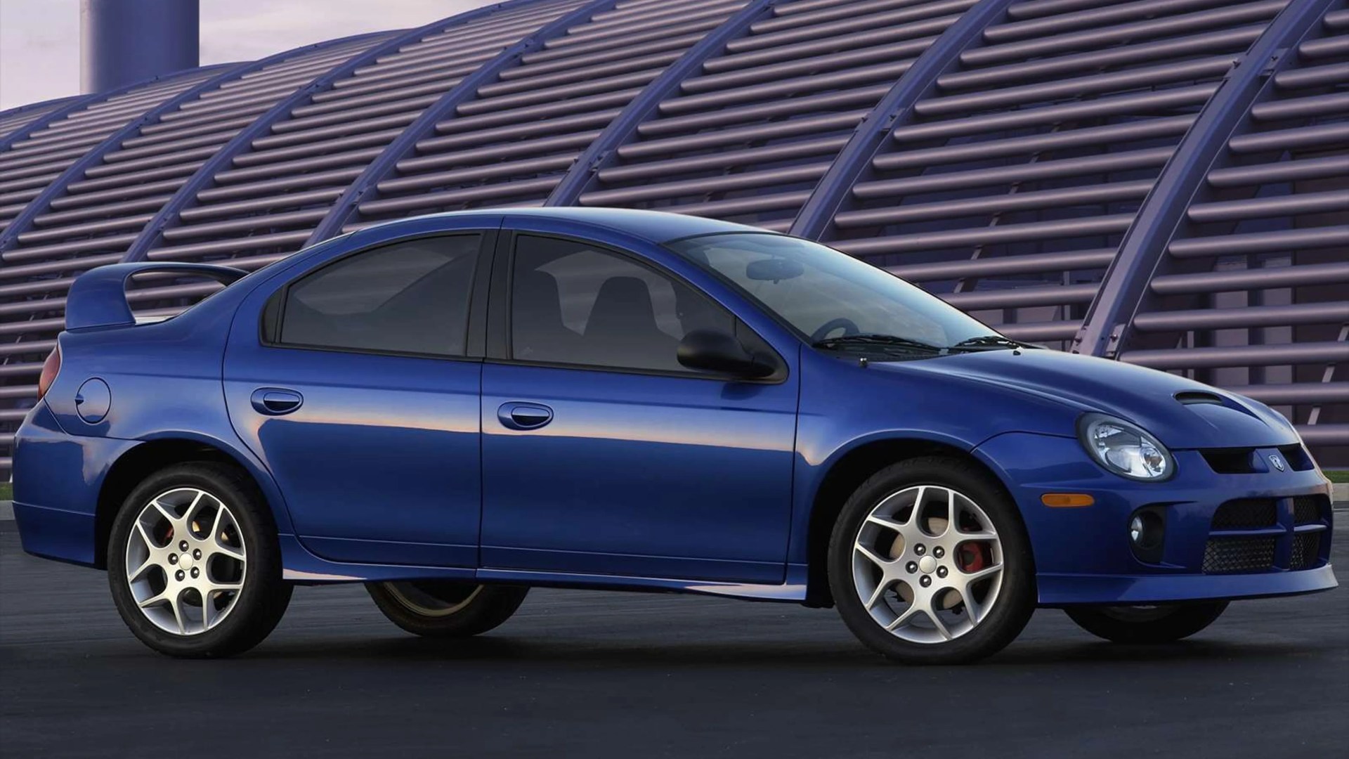 Blue four-door sports sedan with rear spoiler and silver alloy wheels parked in front of a modern architectural structure.
