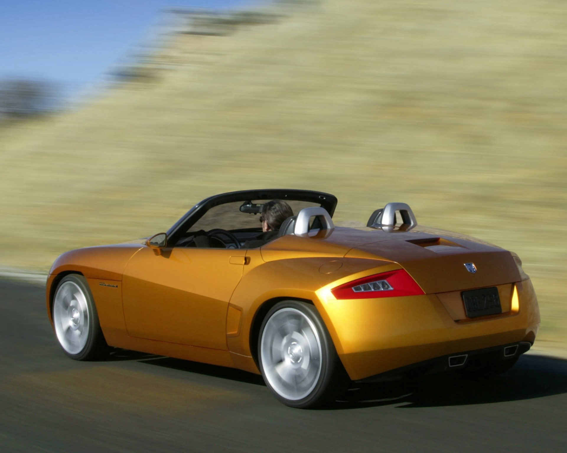 Orange convertible sports car with silver roll bars driving on a road.