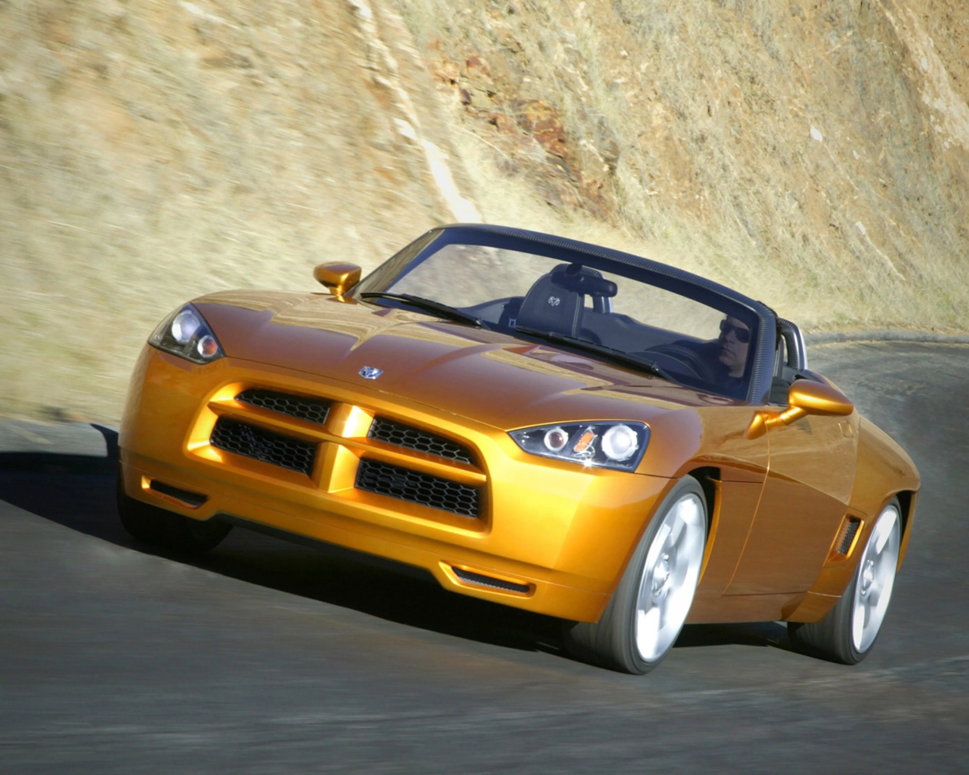 Orange convertible sports car with black interior driving on a curved road near a rocky hillside.