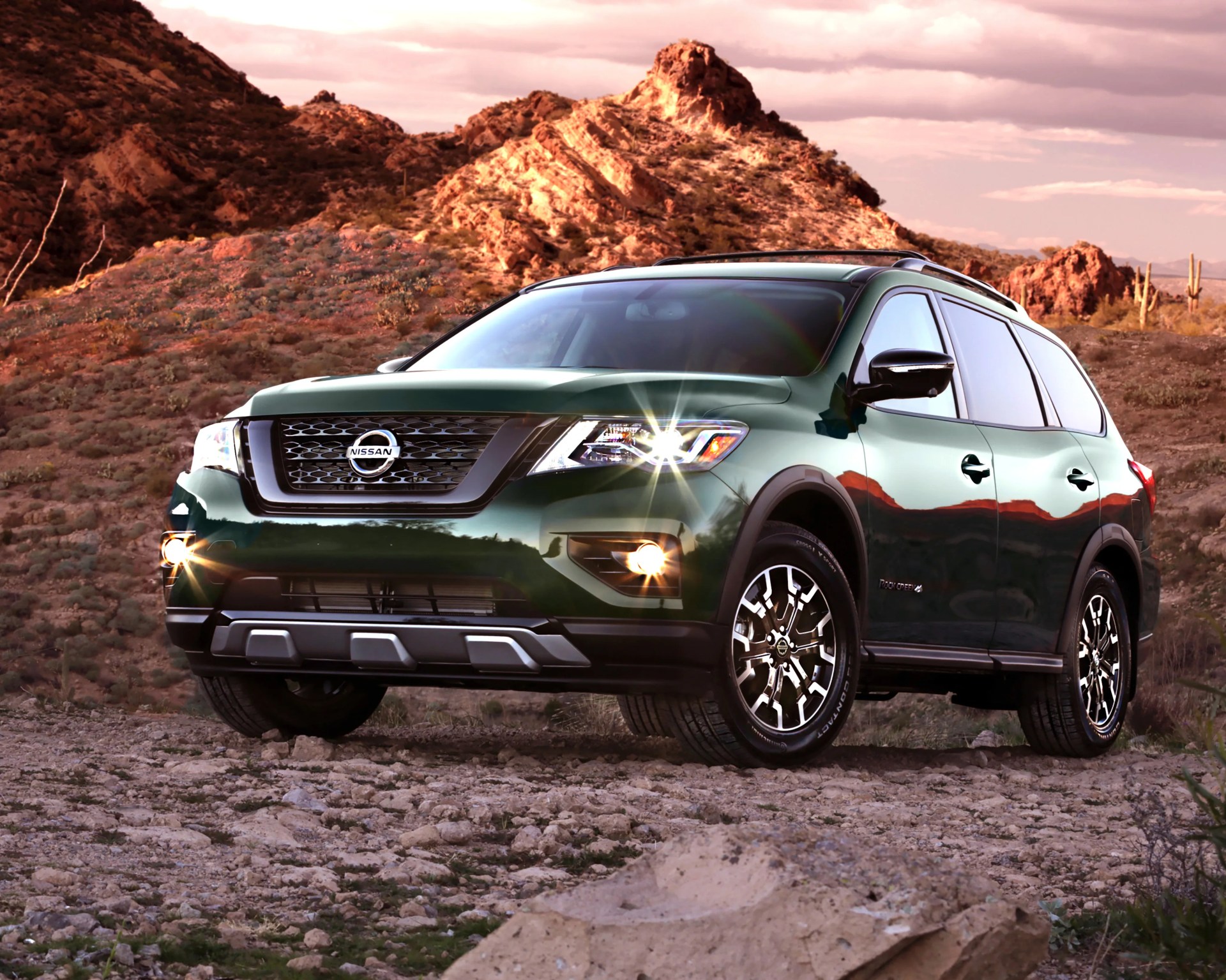 Dark green Nissan SUV with headlights on parked on rocky terrain with desert mountains in the background.