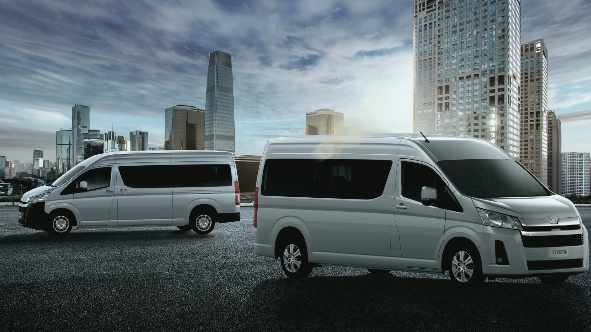 Two silver Toyota vans parked on an asphalt surface with a city skyline and tall buildings in the background.