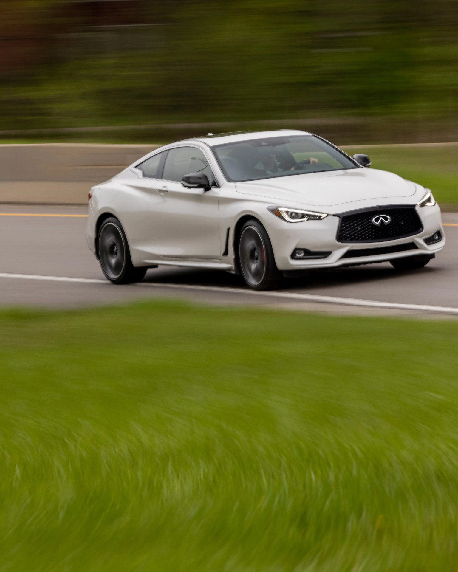 White Infiniti coupe with black grille and wheels driving on a road with green grass in the foreground.