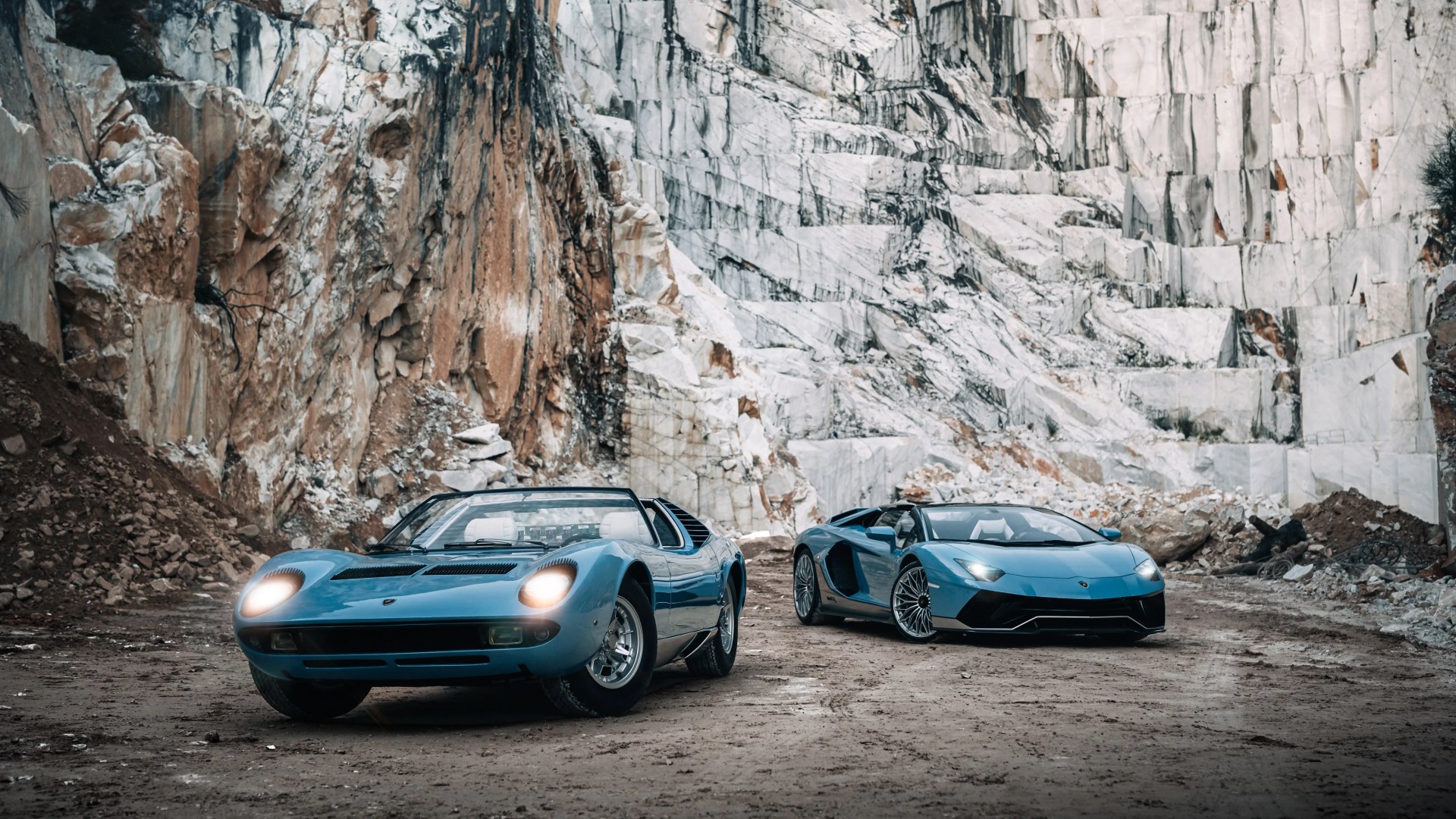 Two blue Lamborghini sports cars, one classic with headlights on and one modern, parked on dirt in a marble quarry.