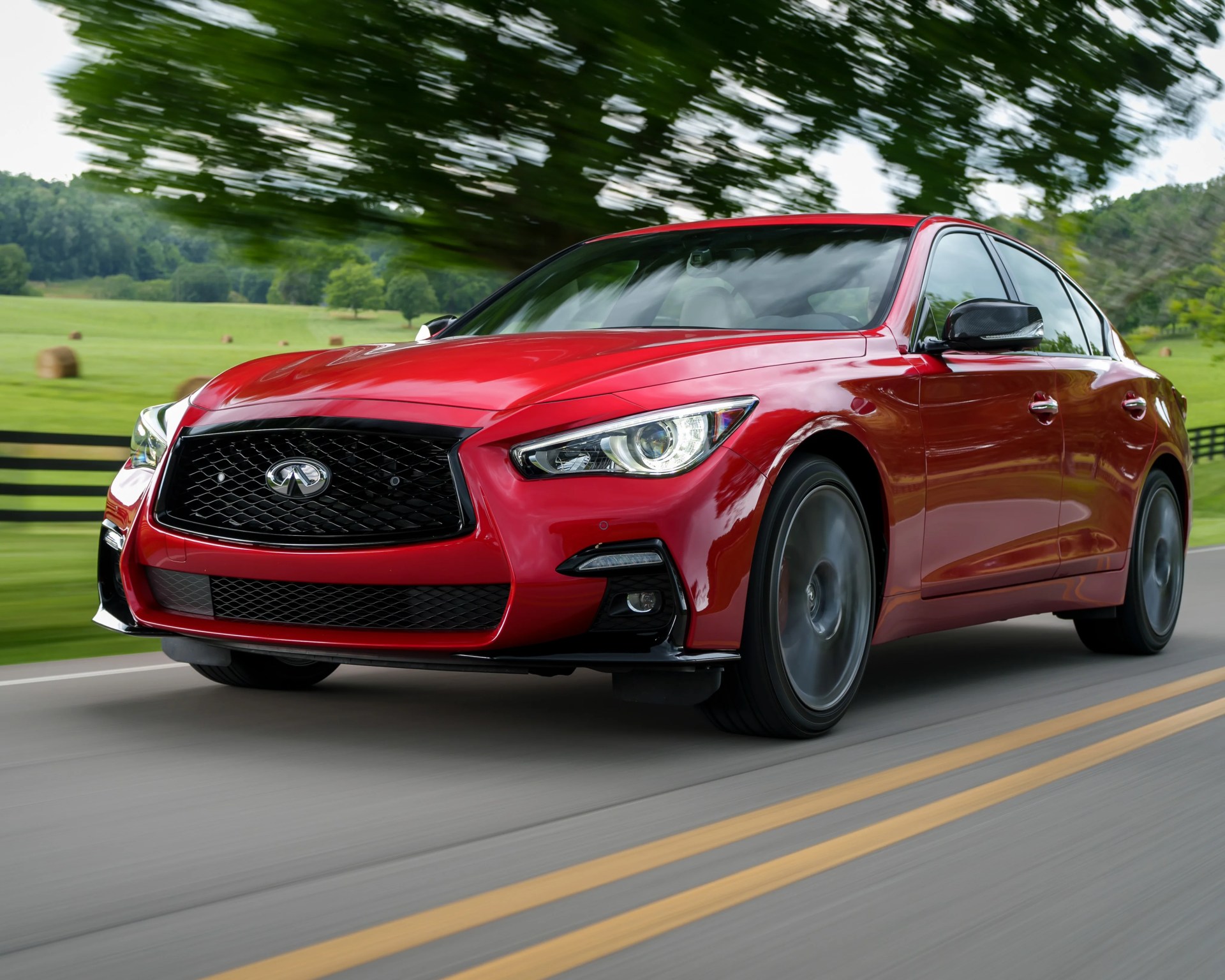 Red Infiniti sedan driving on a road with green fields and trees in the background.