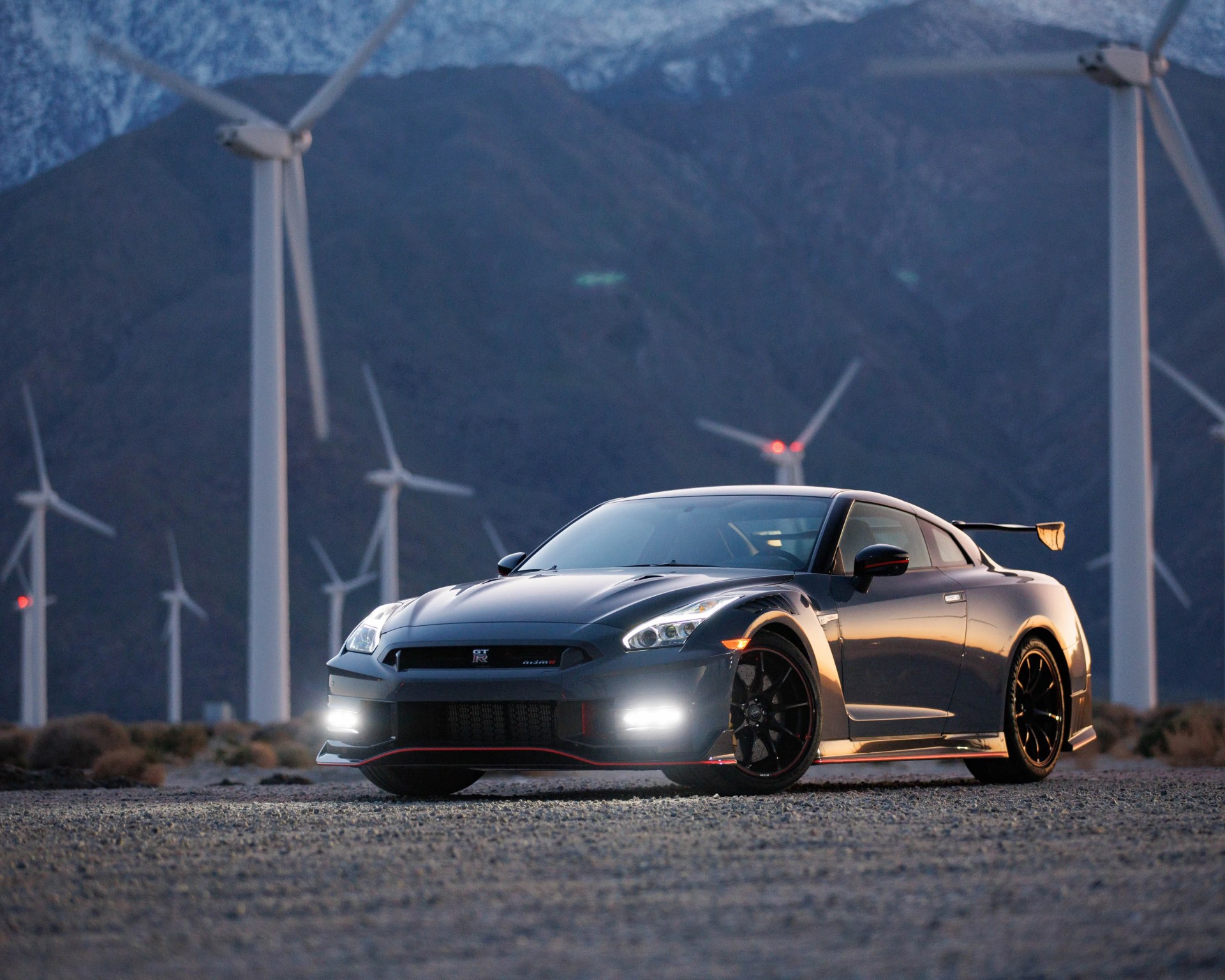 Dark gray Nissan GT-R Nismo sports car with headlights on, parked on gravel with wind turbines and mountains in the background.