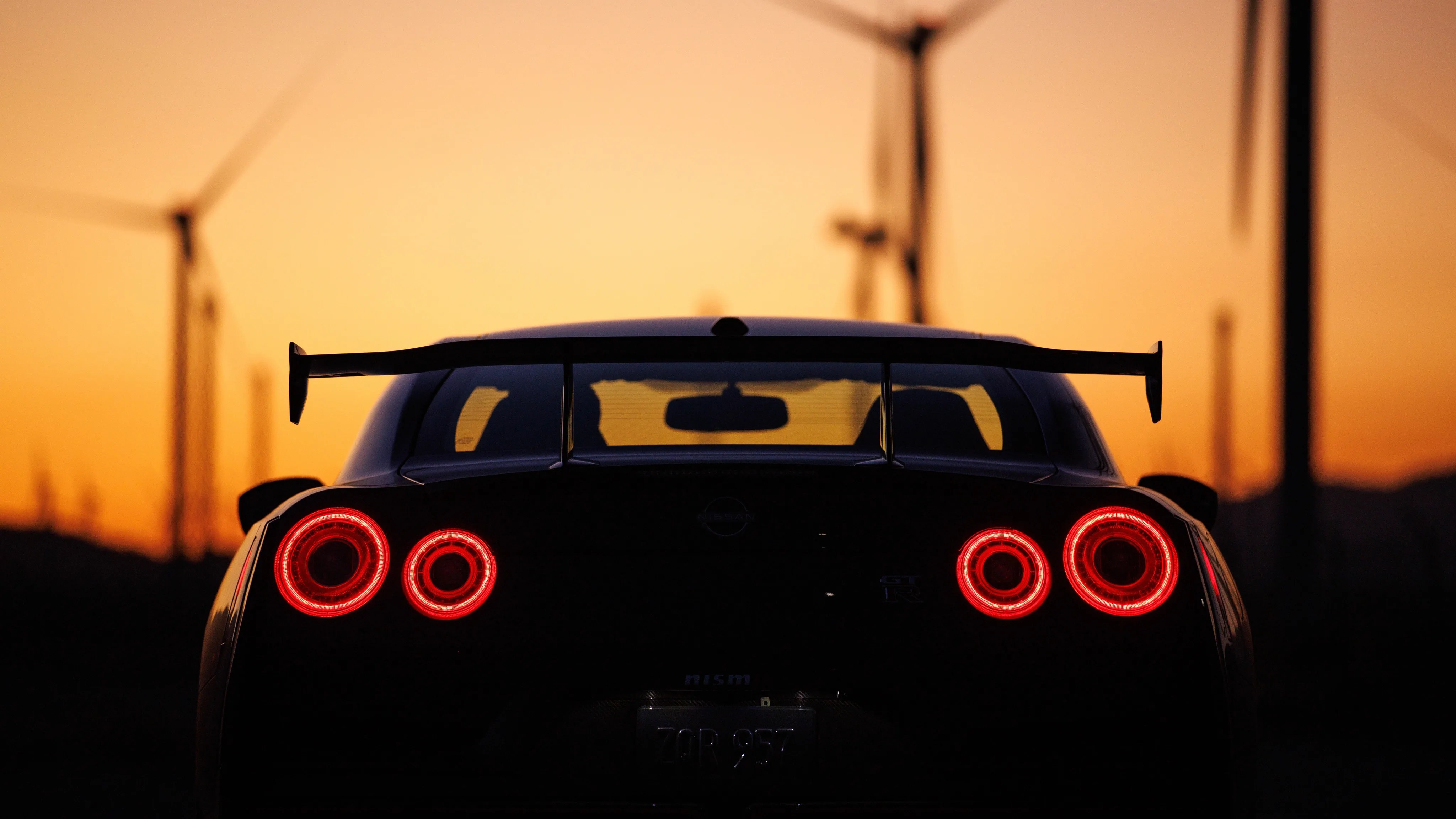 Rear view of a black Nissan GT-R with illuminated round taillights at sunset with wind turbines in the background.