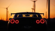 Rear view of a black Nissan GT-R with illuminated round taillights at sunset with wind turbines in the background.