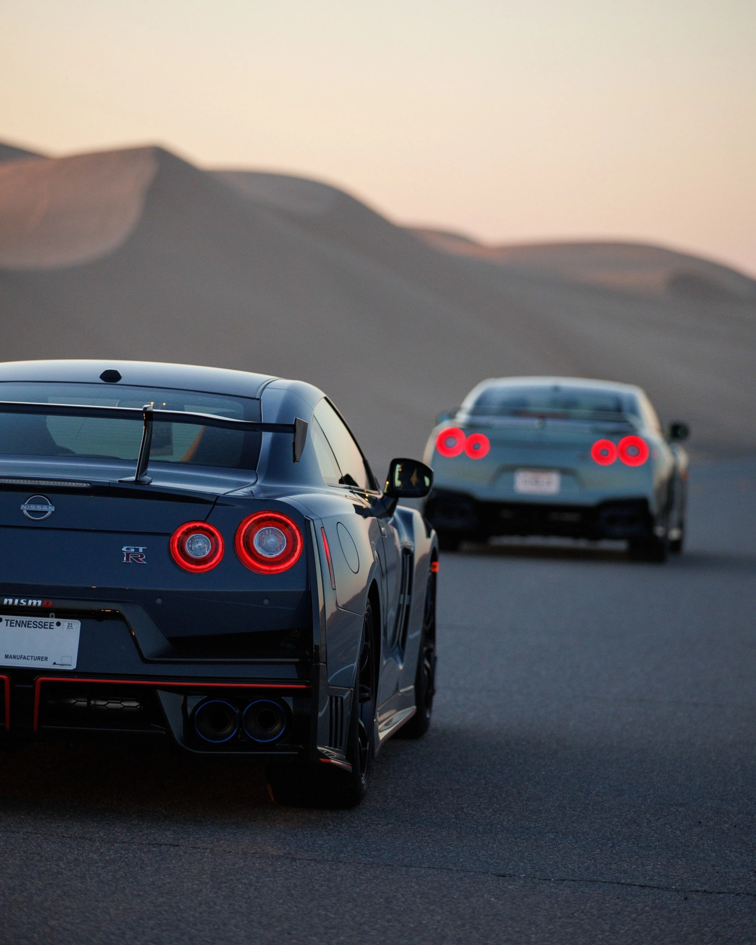 Two Nissan GT-R sports cars with illuminated taillights on a road with sand dunes in the background at dusk.