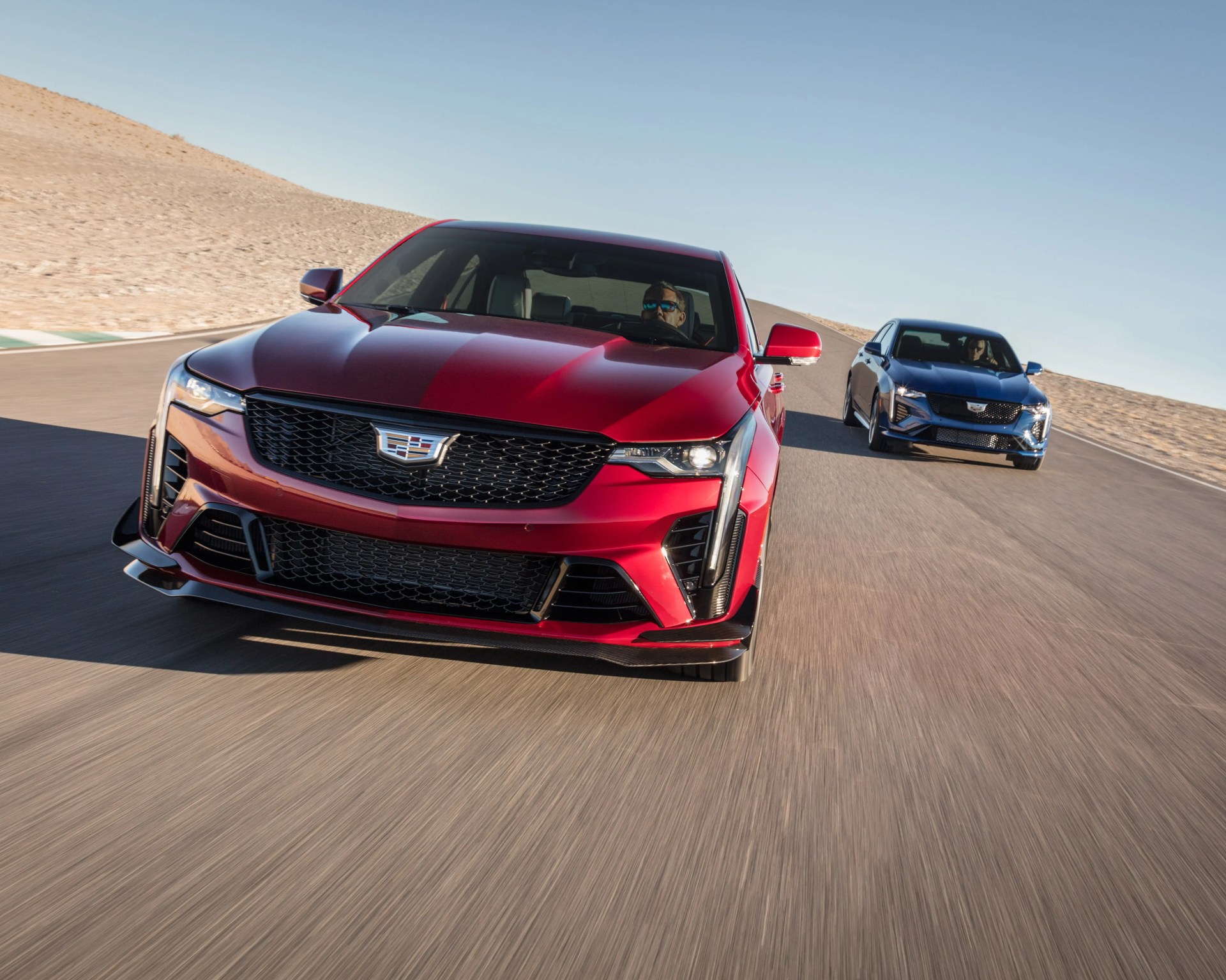 Red and blue Cadillac sedans driving on a desert race track under clear sky.