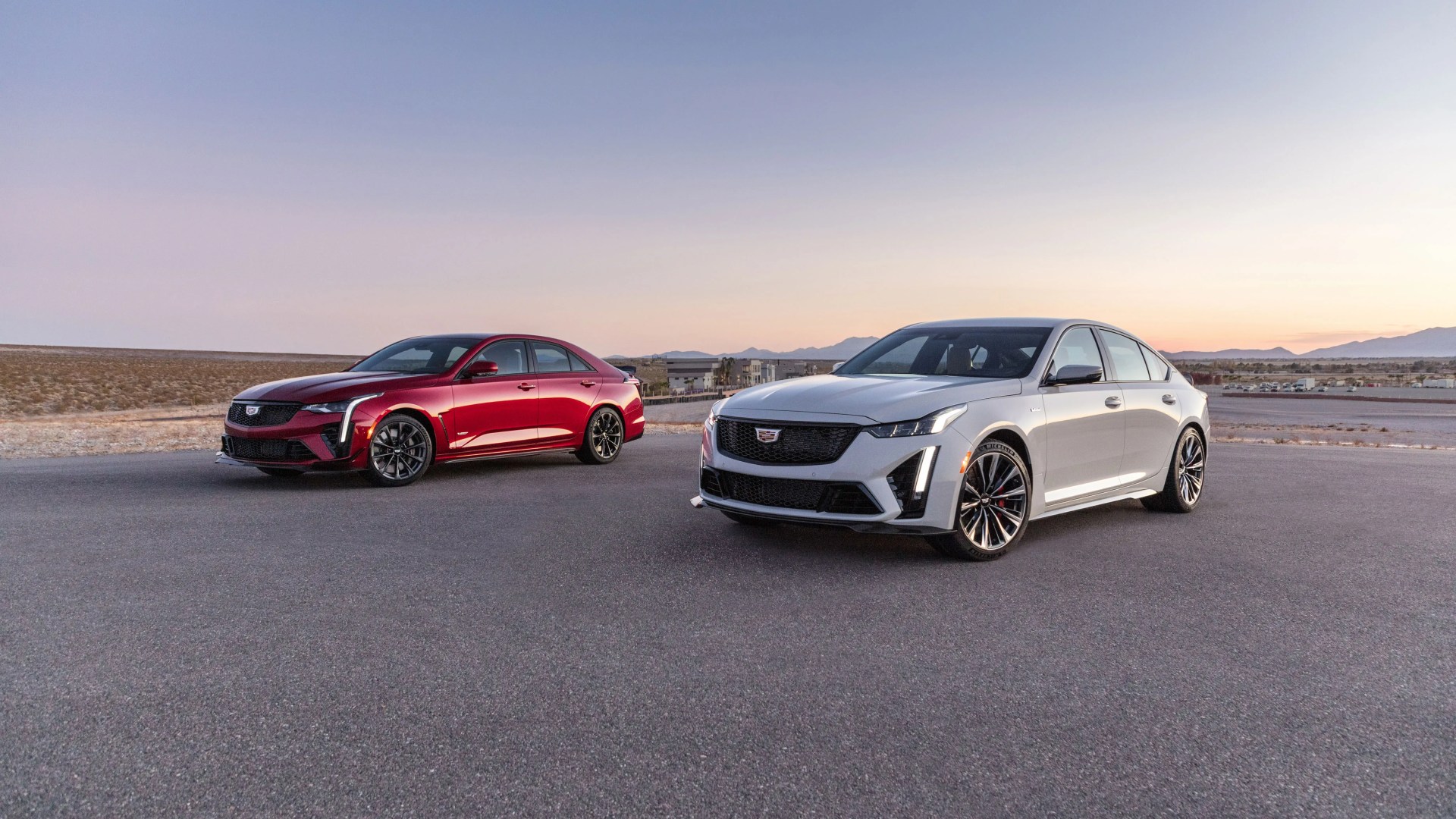 Two Cadillac sedans, one red and one white, parked on an open paved area with mountains in the background at sunset.