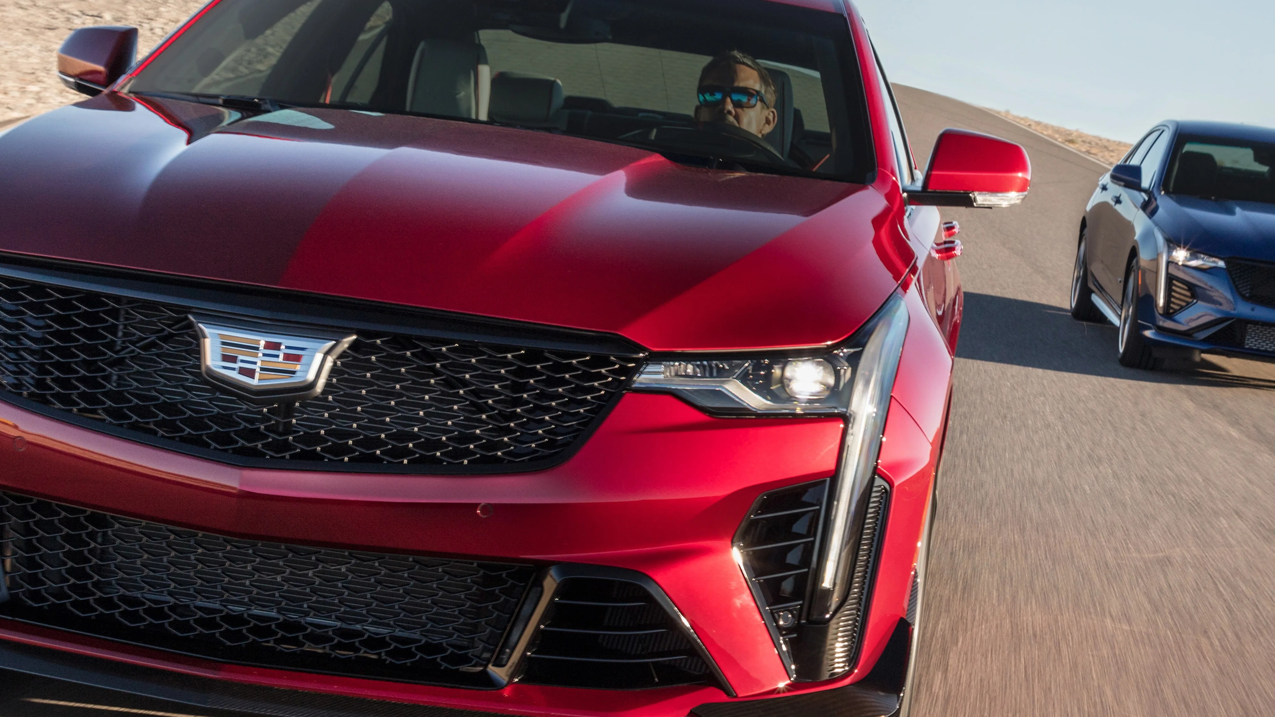 Front view of a red Cadillac sedan with a driver wearing sunglasses, followed by a blue Cadillac on a paved road.