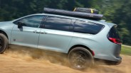 Light blue minivan with black wheels and roof rack carrying a spare tire and covered gear driving on a dirt road.