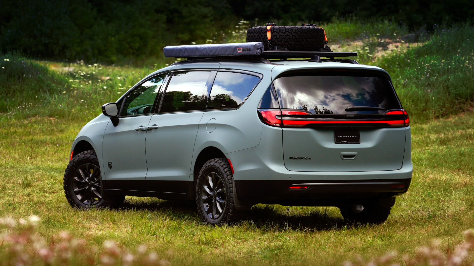 Matte light green Chrysler Pacifica minivan with off-road tires and roof rack carrying a spare tire and covered gear, parked on grass.