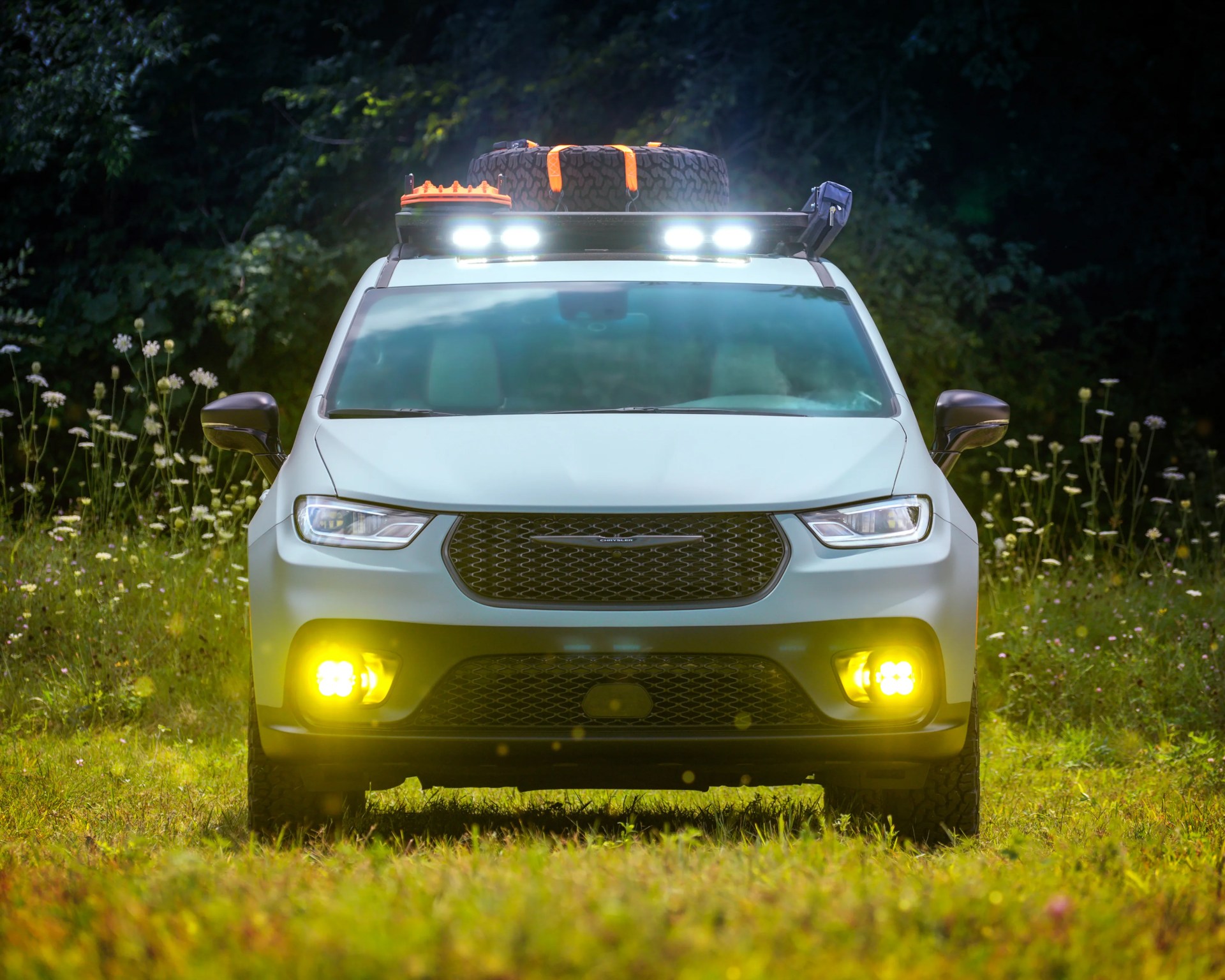 White Chrysler SUV with roof rack carrying a spare tire and gear, headlights and yellow fog lights on in a grassy area.