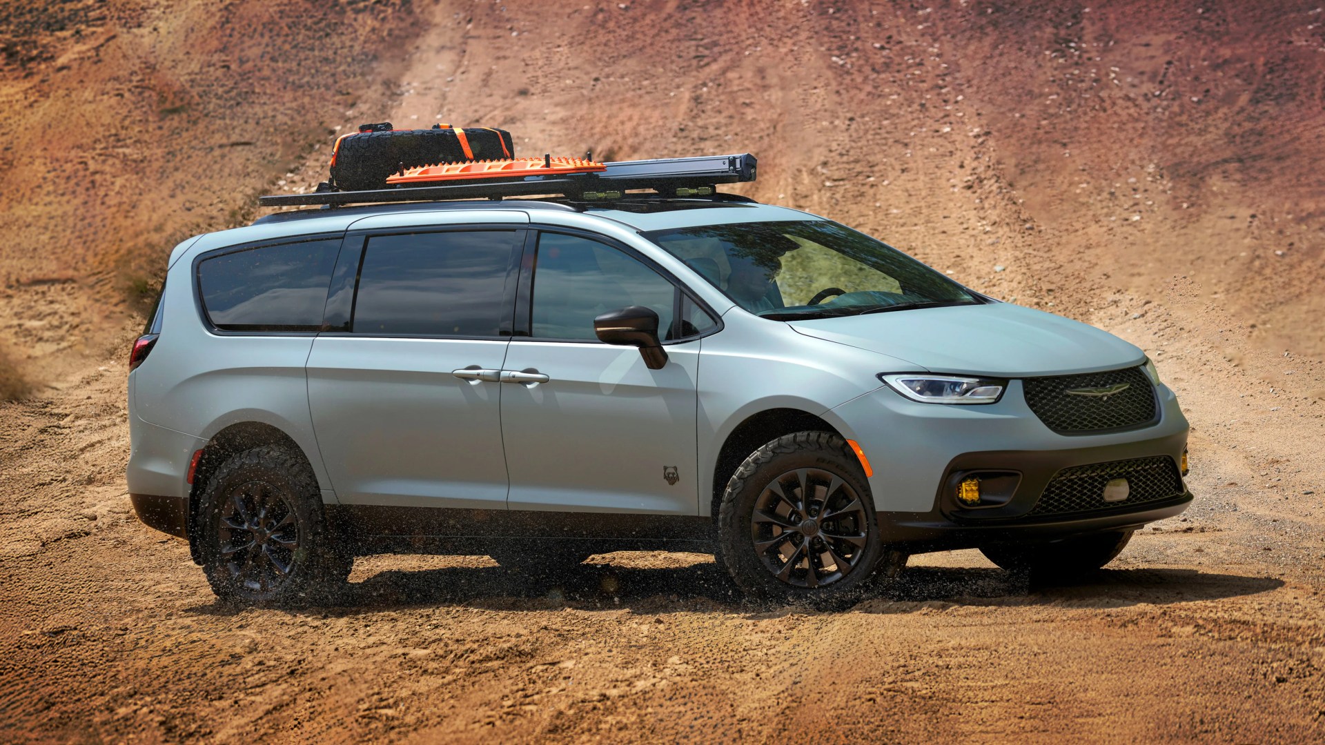 Light gray off-road minivan with black wheels and roof rack carrying a spare tire and orange recovery boards on a dirt trail.