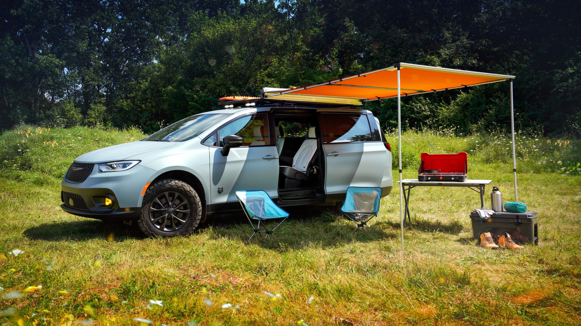 Light blue minivan with roof rack and orange awning extended, two blue camping chairs, folding table with stove, and camping gear on grass.