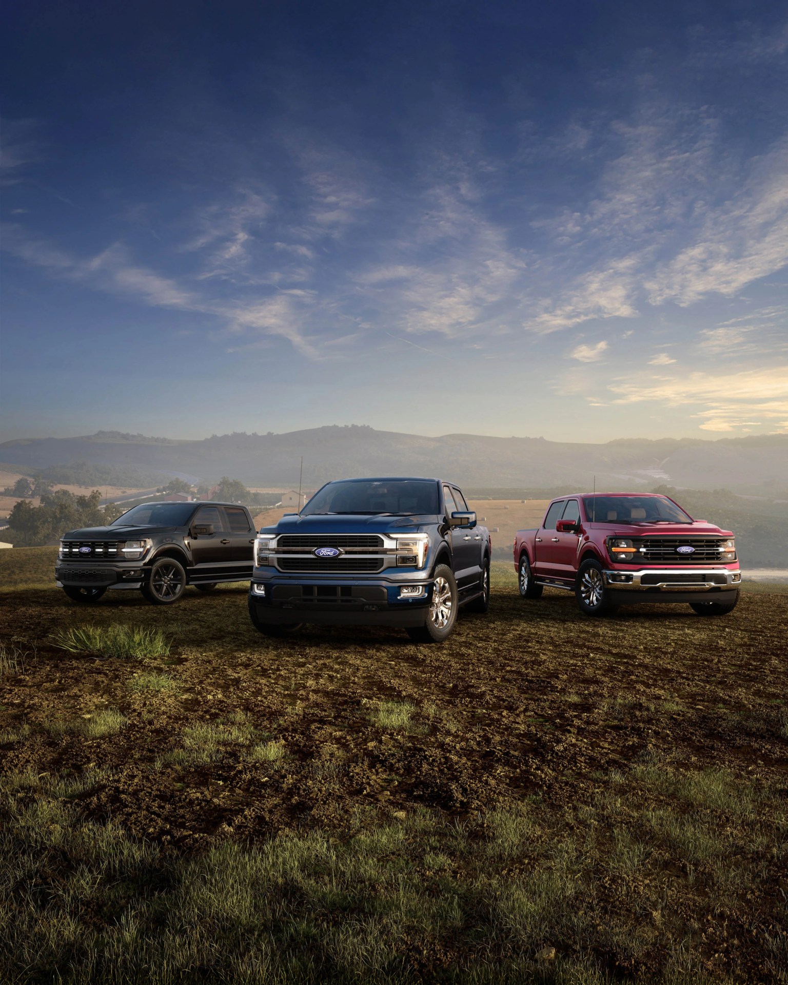 Three Ford pickup trucks in black, dark blue, and red parked on a grassy field with hills and a cloudy sky in the background.